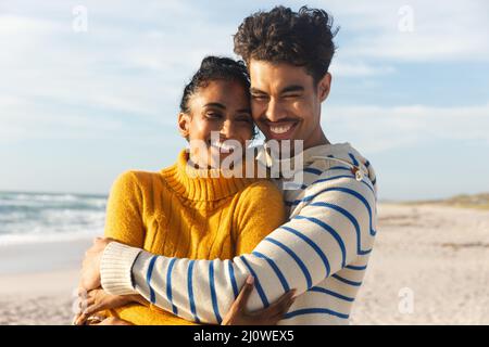 Felice uomo biraciale abbracciando la ragazza da dietro in spiaggia contro il cielo in giorno di sole Foto Stock