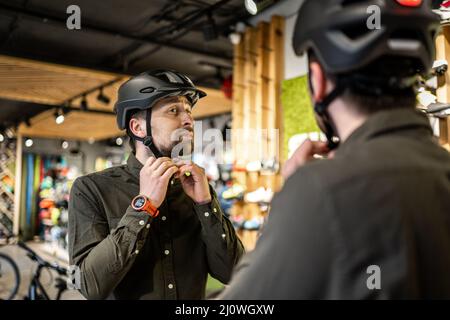 Il giovane uomo è venuto al negozio di biciclette. Sta misurando il casco. Il maschio sceglie il casco nel negozio di attrezzature sportive. Acquisto di nuovo Foto Stock