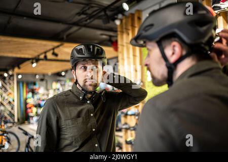 Il giovane uomo è venuto al negozio di biciclette. Sta misurando il casco. Il maschio sceglie il casco nel negozio di attrezzature sportive. Acquisto di nuovo Foto Stock