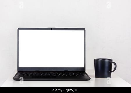 Un mockup bianco sullo schermo di un computer portatile e una tazza nera di tè su un tavolo contro una parete bianca con uno spazio copia Foto Stock