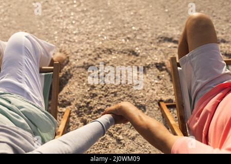 Vista ad angolo alto della coppia multirazziale senior che tiene le mani mentre si rilassa sulle sedie in spiaggia Foto Stock