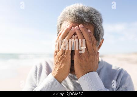 Primo piano di un uomo anziano biraciale preoccupato che copre il viso con le mani in spiaggia contro il cielo nelle giornate di sole Foto Stock