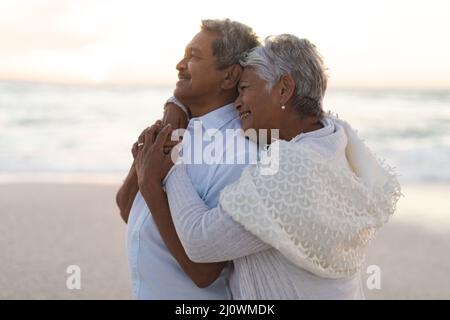 Sorridente coppia multirazziale senior newlywed che guarda via mentre abbraccia la spiaggia durante il tramonto Foto Stock