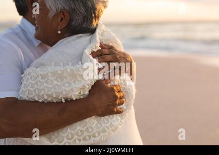 Parte centrale della coppia multirazziale senior newlywed che si abbraccia a vicenda in spiaggia durante il tramonto Foto Stock