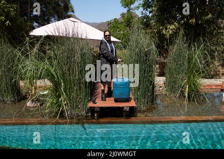 Donna messicana caucasica vestita con un costume da bagno, mantello, stivali e occhiali da sole che arrivano in una piscina naturale all'aperto dirigendo una valigia con ruote su un w Foto Stock