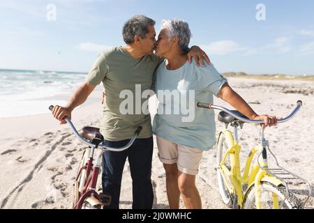 Affettuoso coppia multirazziale anziano baciare mentre in piedi con i cicli a spiaggia soleggiata contro il cielo Foto Stock