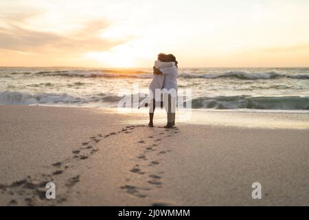 Vista laterale della coppia multirazziale senior newlywed che si abbraccia sulla spiaggia durante il tramonto Foto Stock