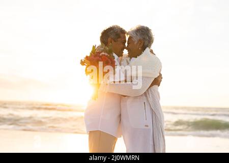 Felice giovane coppia multirazziale senior abbracciando mentre tiene bouquet in spiaggia durante il tramonto Foto Stock