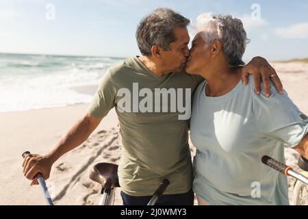 Pensionato multirazziale senior coppia baciare mentre in piedi con biciclette in spiaggia durante la giornata di sole Foto Stock