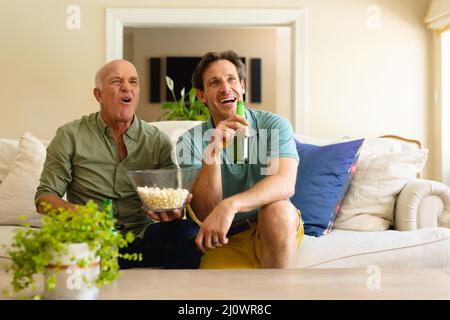 Padre e figlio caucasici con birra e popcorn guardando la tv seduta insieme sul divano a casa Foto Stock