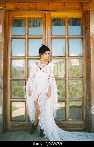La sposa in un abito in vimini bianco è appoggiata contro una porta di vetro Foto Stock