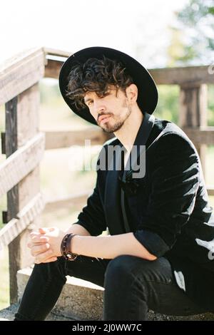 L'uomo in un cappello nero siede sui gradini vicino alla ringhiera di legno Foto Stock