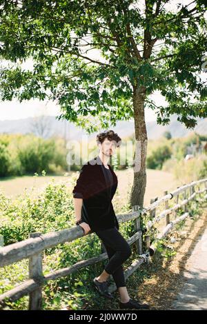 Il giovane uomo in una giacca nera siede su una recinzione di legno vicino ad una strada di campagna Foto Stock