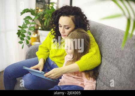 Madre e figlia che guardano i cartoni animati su tablet digitale, seduti sul divano e utilizzando un dispositivo moderno Foto Stock