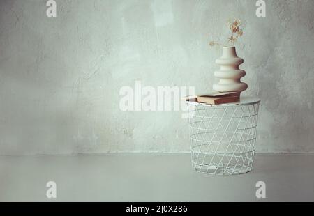 Libro e asciugò ramoscello in vaso di ceramica su comodino rotondo in metallo bianco contro parete di cemento beige Foto Stock