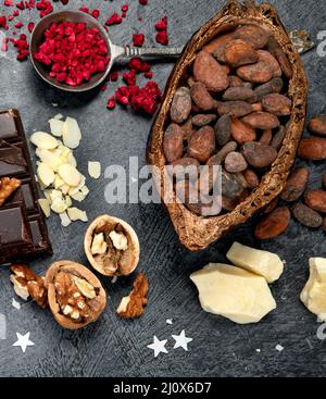 Barra di cioccolato, pezzi di cioccolato fondente schiacciati, fagioli di cacao, burro di cacao, noci, vista dall'alto su sfondo scuro. Foto Stock