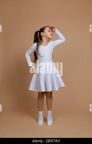 Ritratto di giovane ragazza sorridente carina con lunghi capelli scuri in abito bianco, calzini e scarpe ginniche in piedi sulle dita, guardando via con mano sopra la testa Foto Stock