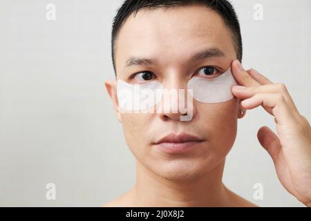 Faccia del giovane uomo asiatico serio che applica il collagene sotto le zone dell'occhio Foto Stock