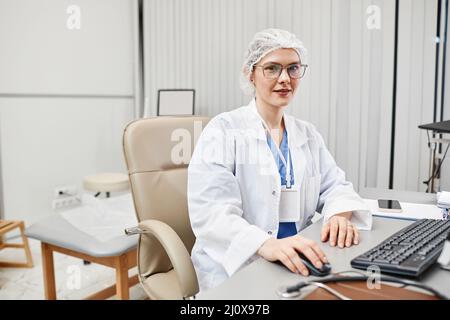Ritratto di medico femminile indossando camice da laboratorio e guardando la macchina fotografica mentre si siede alla scrivania in clinica, spazio copia Foto Stock