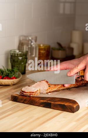 Taglio uomo irriconoscibile con filetto di maiale cotto a coltello grande sul tagliere, verticale Foto Stock