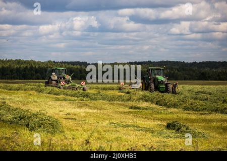 Due trattori John Deere verdi haymaking con aratro Krone sul campo estivo prima della tempesta - teleobiettivo con fuoco selettivo Foto Stock