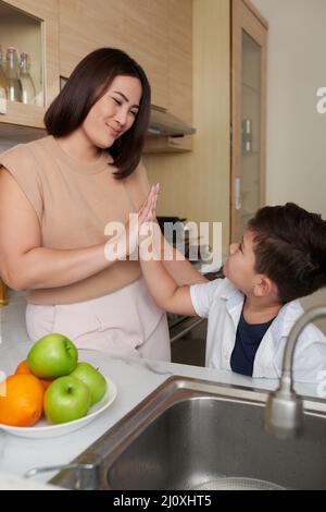 Madre che dà cinque alti al suo figlio piccolo dopo che finiscono per pulire la frutta sotto acqua di rubinetto in cucina Foto Stock