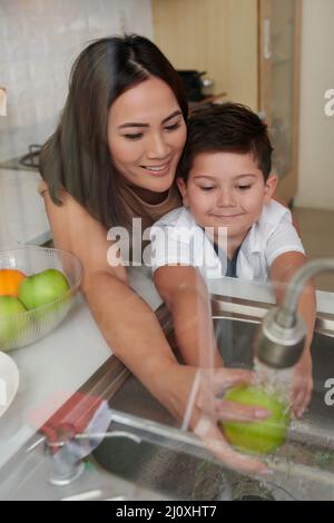 Sorridendo giovane madre che insegna a figliolo di sciacquare la frutta sotto l'acqua prima di mangiare Foto Stock
