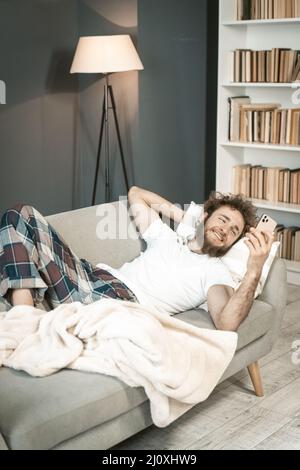 Giovane malato in pigiama che dorme sul divano guardando lo smartphone o facendo una videochiamata . Bel ragazzo bearded che dorme Foto Stock