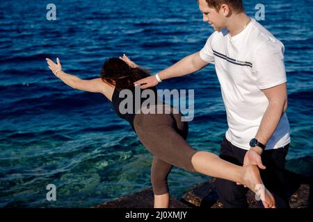 Personal trainer uomo correggere cliente donna mentre fare stretching o yoga all'aperto sfondo mare. Supporto dell'istruttore Concept asana. Foto Stock