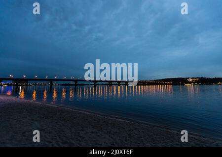 Un ponte sul fiume, edifici illuminati e lanterne sulla riva del fiume, luci colorate della città notturna riflessa nell'acqua Foto Stock