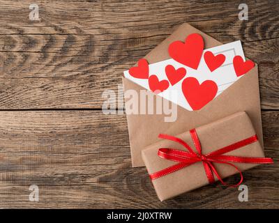 Cartoncino e cuori rossi in busta aperta di carta Kraft marrone. Confezione regalo con nastro rosso su sfondo vintage invecchiato in legno. Greetin Foto Stock