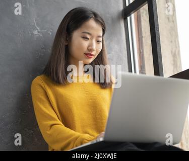 Donna asiatica che lavora su un computer portatile a casa o in un bar sulla finestra. Foto Stock