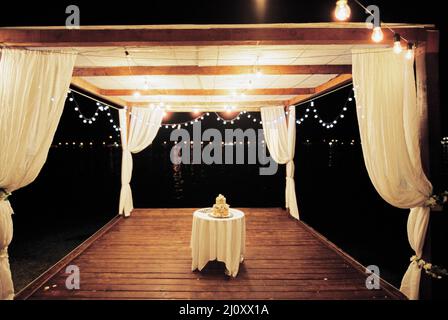 Torta nuziale si trova su un tavolo in un gazebo decorato con lanterne sulla spiaggia Foto Stock