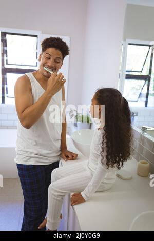 L'uomo biraciale allegro spazzolando i denti mentre guarda la figlia seduta sul banco del bagno Foto Stock