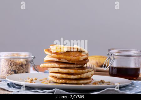 Pancake con fette di banana e sciroppo serviti in piatto da avena e vaso di miele su sfondo grigio Foto Stock