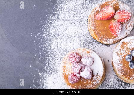 Vista dall'alto dello zucchero in polvere sopra le frittelle accatastate con vari frutti di bosco su ardesia alimentare Foto Stock