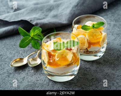 Dessert a strati sani con yogurt, banana, marmellata di mango, biscotti in vetro su sfondo di pietra grigia, vista laterale Foto Stock