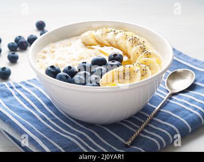 Fiocchi d'avena con banane, mirtilli, chia, marmellata, miele, tovagliolo blu su sfondo bianco in legno, vista laterale Foto Stock