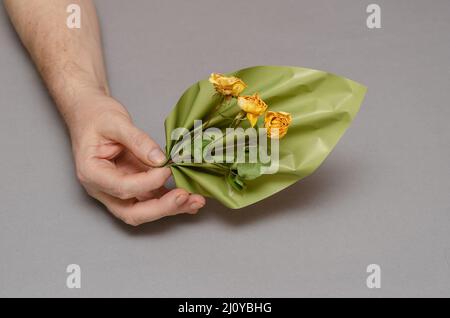 Due mani maschili tengono un elemento decorativo ondulato e fiori gialli su uno sfondo grigio. Una figura di plastica verde a forma di foglia di pianta. Tre drie Foto Stock