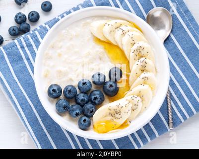 Fiocchi d'avena con banane, mirtilli, chia, marmellata, miele, tovagliolo blu su sfondo bianco in legno. Primo piano. Vista dall'alto Foto Stock