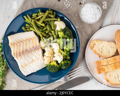 Cotto pesci di mare filetti di merluzzo con verdure sulla piastra blu, pane, grigio igienico, sullo sfondo di legno, vista dall'alto. Una corretta dieta Foto Stock