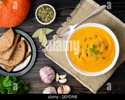 Zuppa di zucca schiacciata in un piatto bianco, semi di zucca, pane, aglio su sfondo di legno scuro Foto Stock