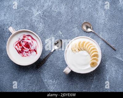 Due yogurt greco con marmellata e banana in ciotola bianca su un tavolo grigio in cemento blu, vista dall'alto Foto Stock