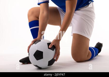 Sezione bassa di giovane donna biraciale che tiene la palla di calcio mentre si inginocchiano su sfondo bianco Foto Stock