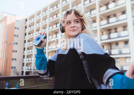 90s estetica. Scatto all'aperto su un blocco di appartamenti. Ritratto di allegra e positiva giovane caucasica che ascolta musica sul suo lettore di cassette. Foto di alta qualità Foto Stock