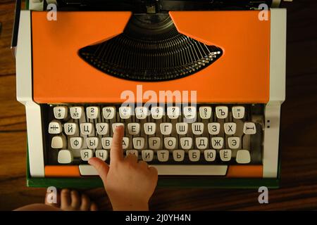 La mano dei bambini preme la lettera russa sulla macchina da scrivere arancione vintage. Il bambino impara le lettere. Foto Stock