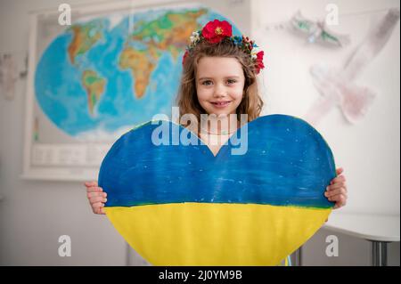 Piccola studentessa Ucraina che tiene il cuore in colori ucraini e guarda la macchina fotografica a scuola. Foto Stock