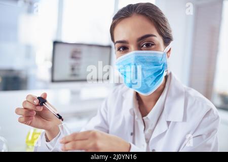 Tempo per alcuni test. Ritratto di una giovane scienziata sicura indossando una maschera chirurgica mentre si fa un esperimento e guardando la macchina fotografica Foto Stock