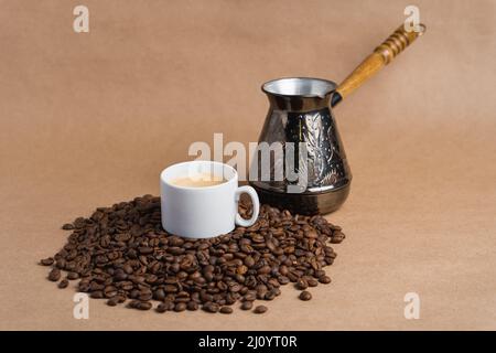 Piccola tazza di caffè bianca con crema espresso e cezve su fagioli tostati su sfondo marrone. Mock up Foto Stock