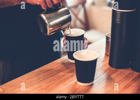 Tagliare il barista versando il latte caldo dalla tazza di ferro in tazze di caffè. Alto angolo di anonimo professionista che fa cappuccino e latte da portare via Foto Stock